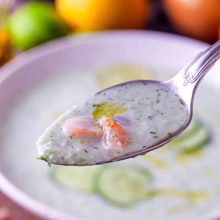 Spoon held over a white bowl showing a scoop of creamy chilled cucumber avocado soup with an optional shrimp piece and cucumber slices visible in the bowl below.