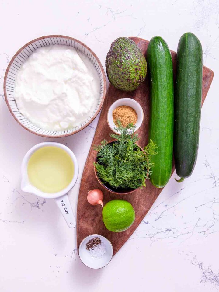 Flat lay of ingredients for chilled cucumber avocado soup including Greek yogurt, two English cucumbers, avocado, fresh dill, shallot, lime, ground cumin, vegetable broth, salt and pepper.