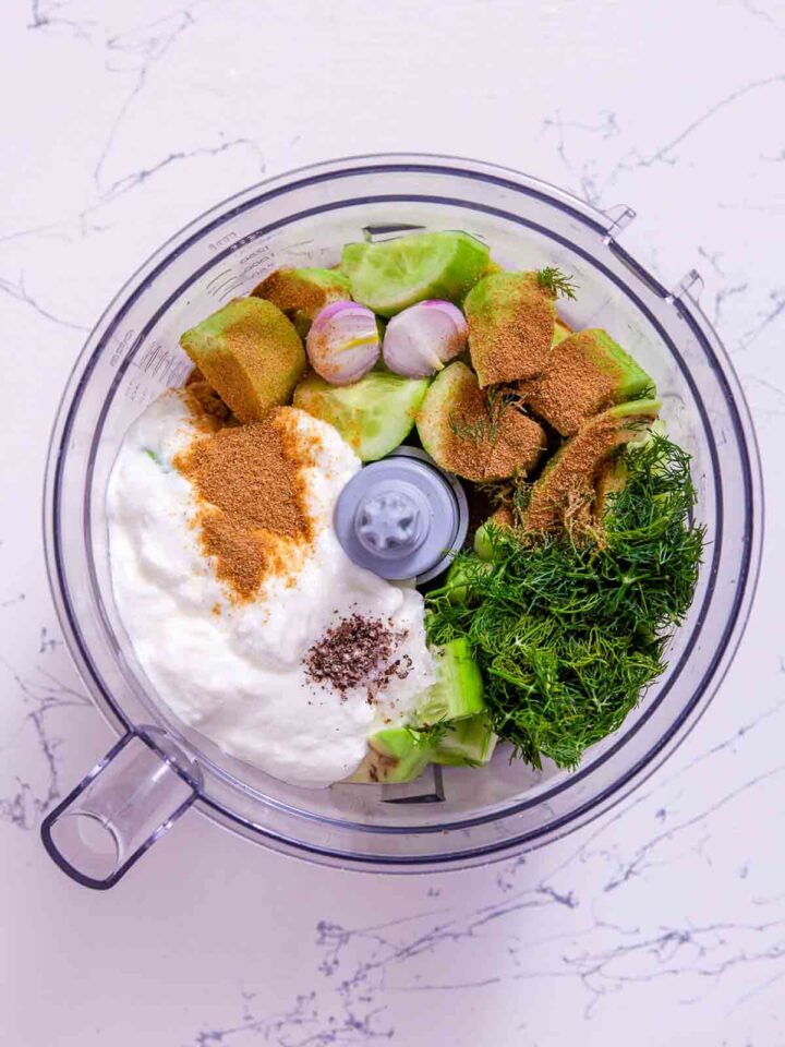 Food processor bowl filled with cucumber chunks, avocado pieces, shallot halves, Greek yogurt, fresh dill, and ground cumin before blending.