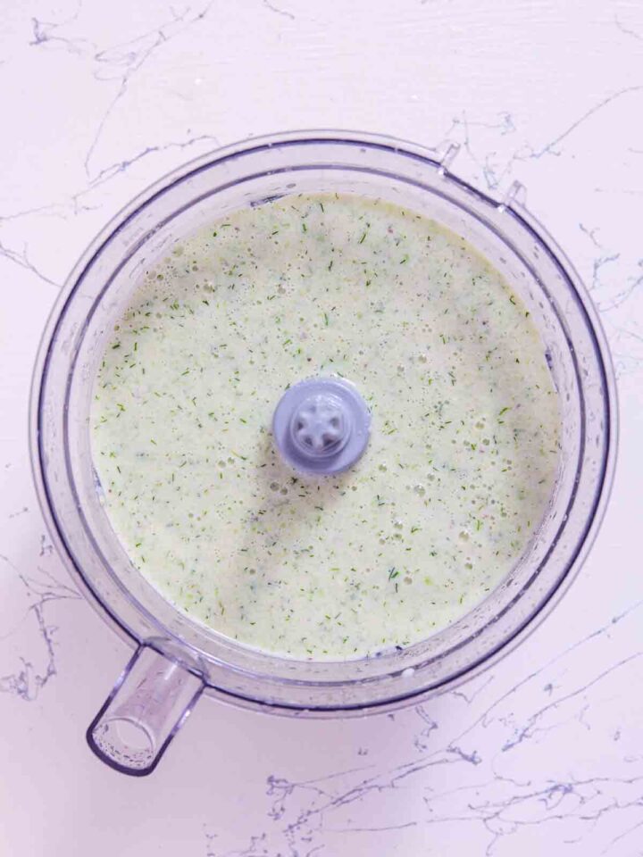 Blended cucumber avocado soup in a food processor, pale and creamy with visible green dill flecks, smooth and ready to chill.