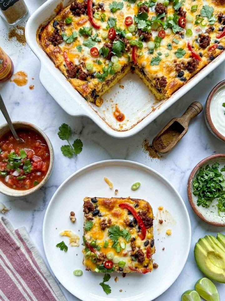 Overhead shot of a slice of Mexican breakfast casserole with chorizo, black beans, red bell pepper, and melted cheese on a white plate, with the baking dish in the background.