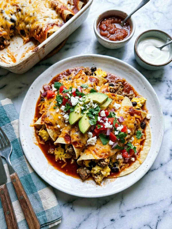 Overhead shot of Mexican enchilada breakfast casserole on a speckled ceramic plate, showing corn tortilla enchiladas smothered in red enchilada sauce and melted cheese, filled with scrambled eggs, ground chorizo, and black beans, topped with sliced avocado, diced tomato, red onion, crumbled cotija, and fresh cilantro, with the baking dish, salsa, and sour cream in the background.