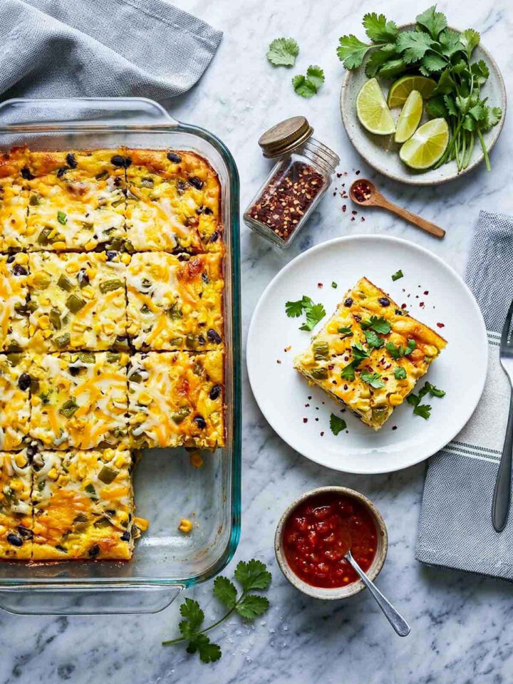 Overhead shot of a make-ahead Mexican egg bake in a glass dish, pre-sliced into squares showing green chiles, roasted corn, black beans, and melted cheese, with one slice served on a white plate garnished with cilantro and chili flakes, and a bowl of salsa on the side.