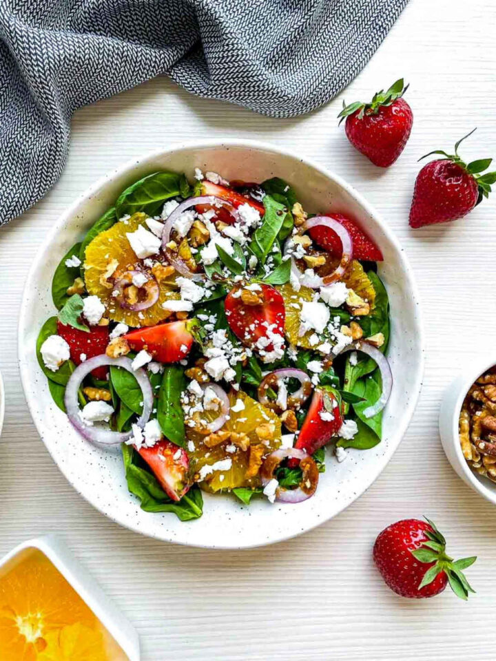Overhead view of strawberry spinach salad with fresh spinach, sliced strawberries, orange segments, red onion, feta cheese, candied walnuts, and balsamic dressing in a white bowl on a light wood surface.