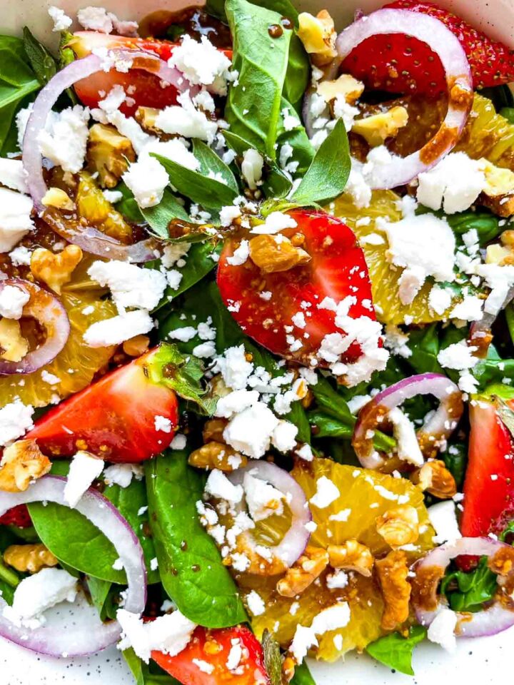 Detailed close-up of strawberry spinach salad with juicy strawberries, orange segments, goat crumbles, walnuts, red onion, and balsamic dressing layered over baby spinach.