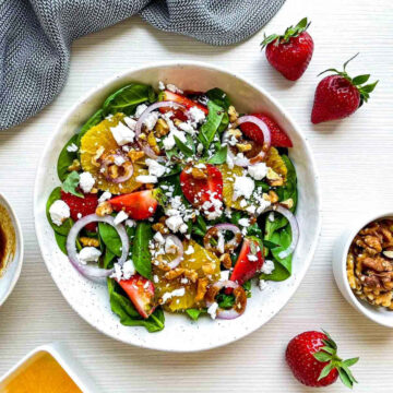 Top-down view of strawberry spinach salad served in a white speckled bowl with goat cheese, strawberries, oranges, walnuts, red onion, and balsamic vinaigrette on a light tabletop.