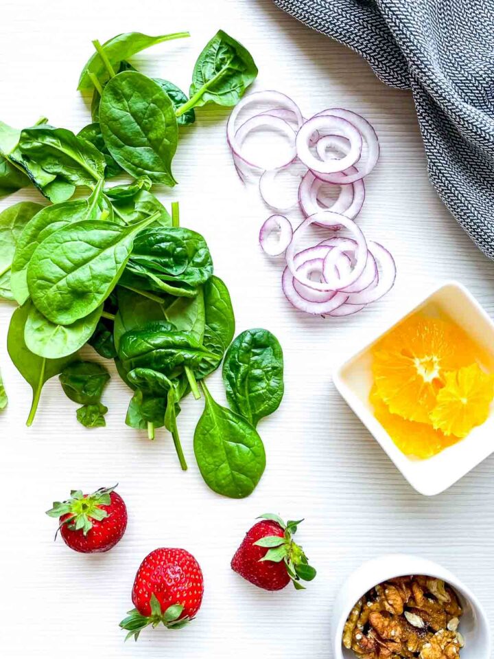 Ingredients for strawberry spinach salad arranged on a light surface with fresh spinach, sliced red onion, orange segments, strawberries, walnuts, and a gray textured napkin.