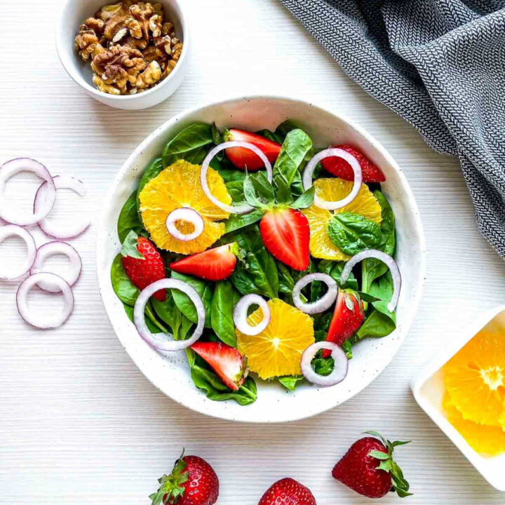 Step-by-step image showing spinach, strawberries, orange slices, and red onion layered in a white bowl for strawberry spinach salad preparation.