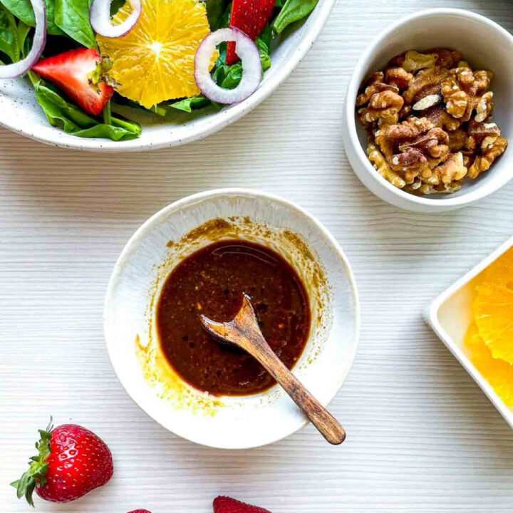 Small bowl of balsamic dressing with a wooden spoon beside ingredients for strawberry spinach salad, including walnuts, orange slices, and fresh strawberries.