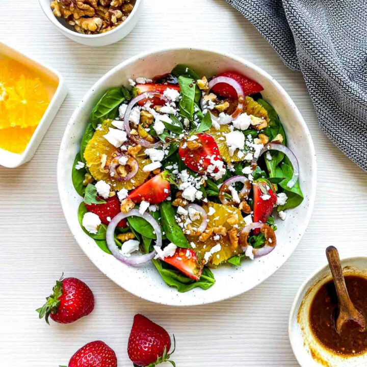 Finished strawberry spinach salad topped with goat cheese, walnuts, red onion, strawberries, orange slices, and balsamic dressing in a white bowl.