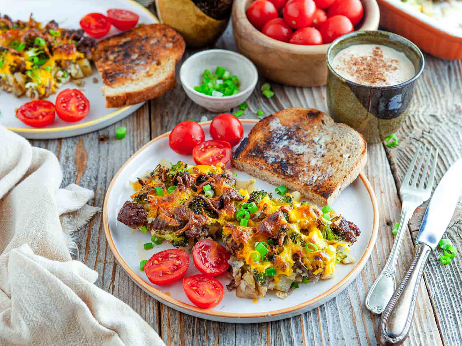 A close angle on a plate of potato sausage broccoli breakfast casserole with toast and cherry tomatoes, coffee mugs and a bowl of tomatoes behind it.