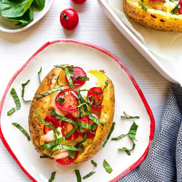 Closeup of a caprese baked potato on a red-rimmed plate, surrounded by fresh basil leaves, whole cherry tomatoes, and a striped napkin, with a second potato in a baking dish nearby.