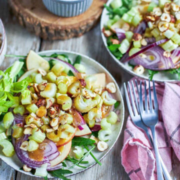 Two plates of apple celery salad topped with toasted hazelnuts, sliced red apple, celery, and red onion on a rustic wood table