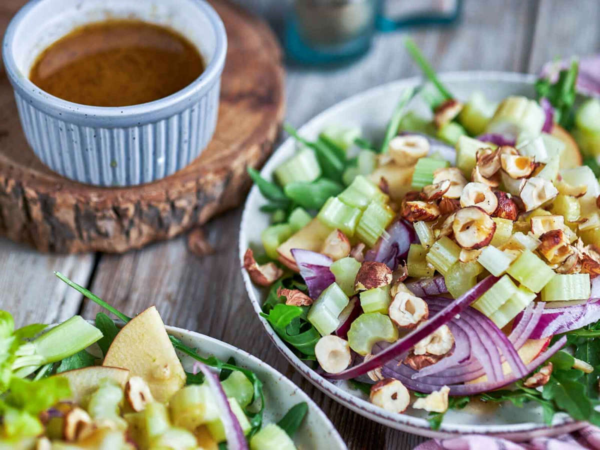 A plated salad of apple, celery, and hazelnuts next to a small jar of vinaigrette on a wood board