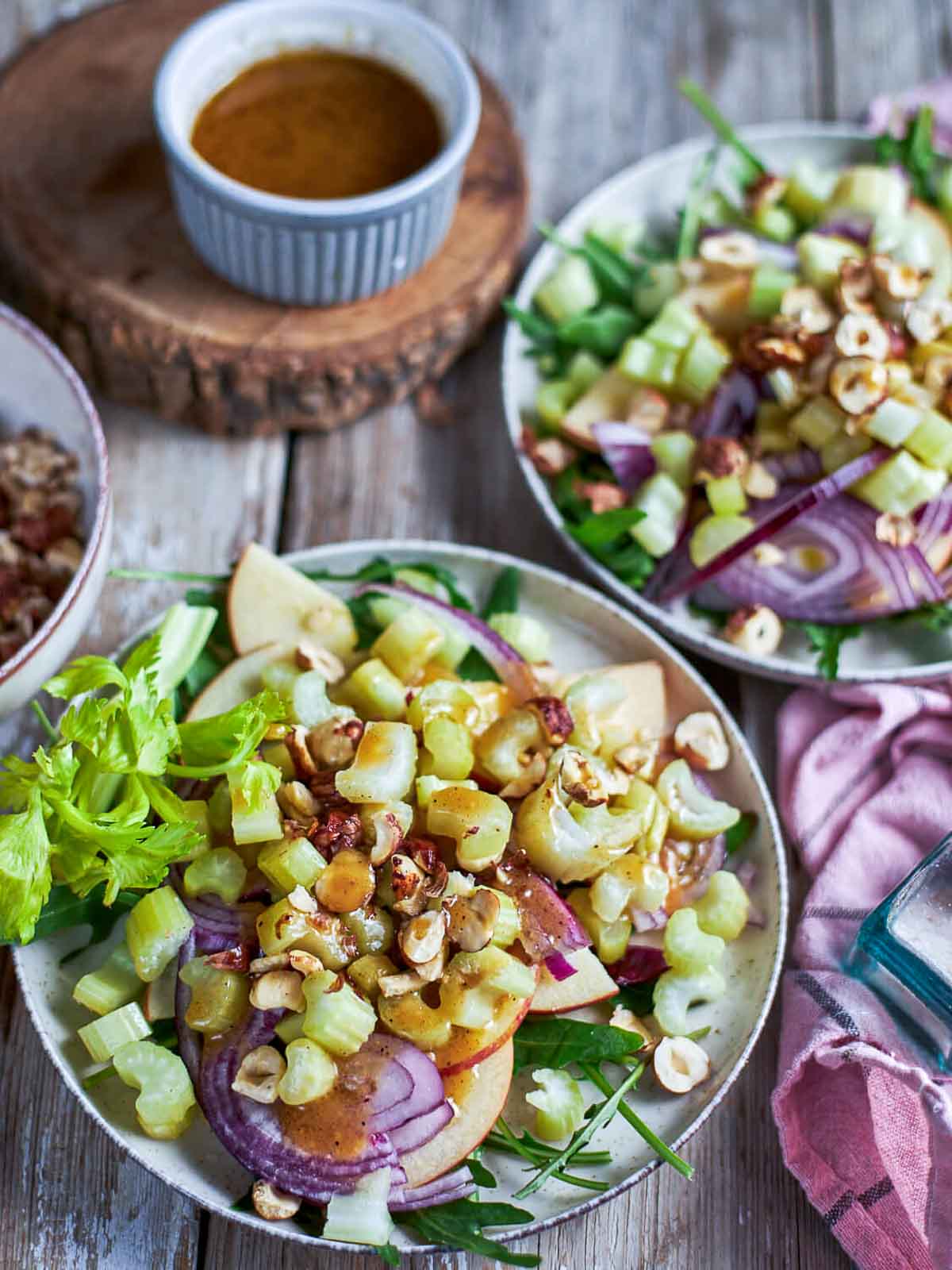 Two plates of dressed salad with visible celery, apple, red onion, and hazelnuts, viewed from above.