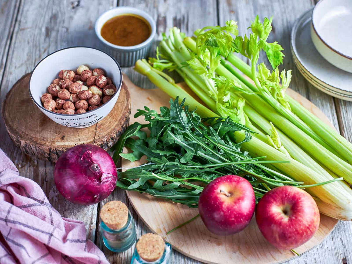 Raw ingredients for the salad arranged on a wood board: whole hazelnuts, celery stalks, arugula, red onion, red apples, and small corked jars of seasoning