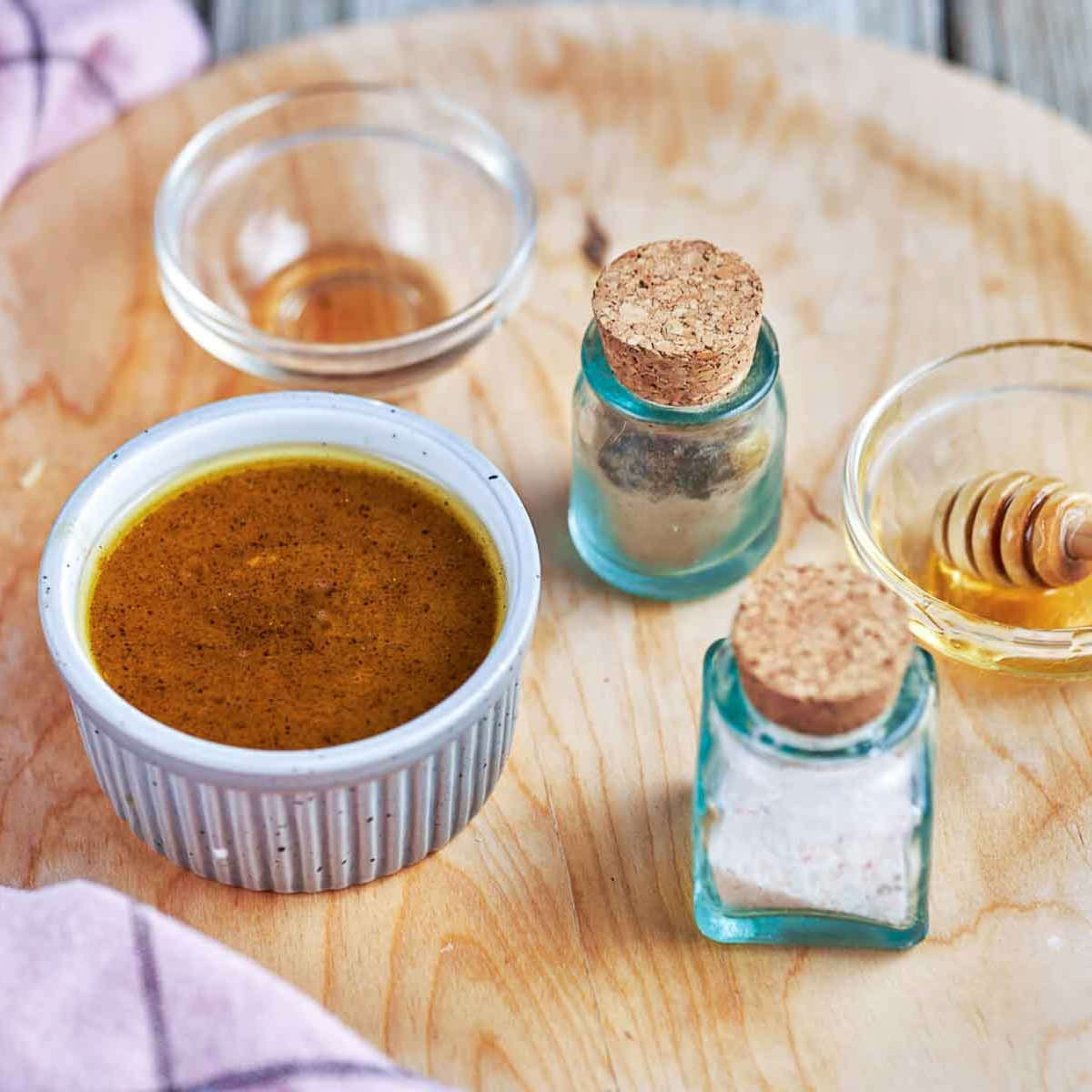 Honey-Dijon vinaigrette in a small ridged ramekin next to bowls of honey and oil and two corked glass jars of seasoning.