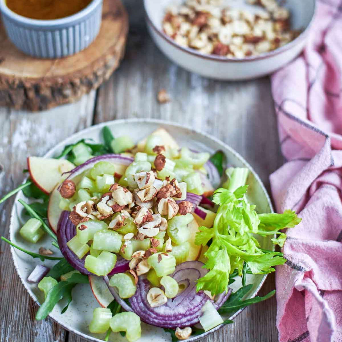 Plated salad topped with chopped hazelnuts, celery, red onion, and apple slices before the vinaigrette is added