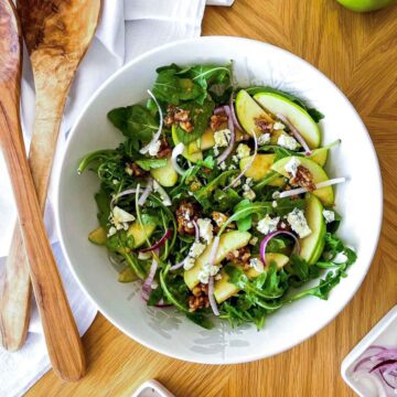 Overhead shot of apple walnut salad with white balsamic vinaigrette, showing arugula, sliced green apples, blue cheese crumbles, candied walnuts, and red onion in a white bowl with wooden salad servers alongside.