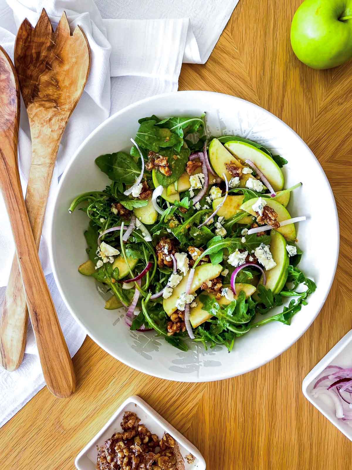 Overhead shot of apple walnut salad with white balsamic vinaigrette, showing arugula, sliced green apples, blue cheese crumbles, candied walnuts, and red onion in a white bowl with wooden salad servers alongside.
