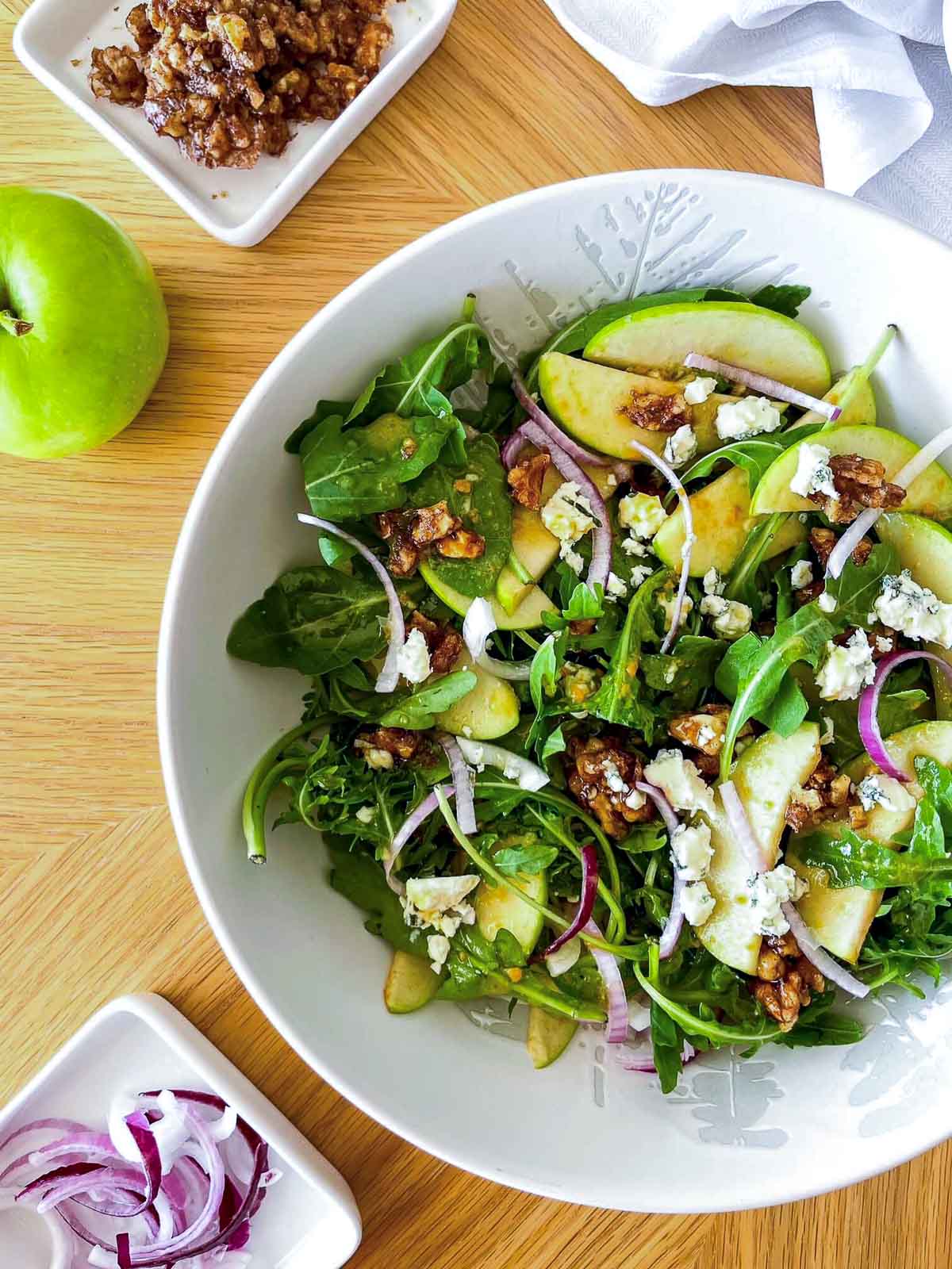Bowl of arugula salad with sliced green apples, blue cheese, candied walnuts, and red onion strands, seen from above next to a whole green apple.