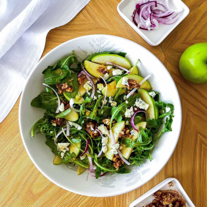 Close overhead view of a tossed green salad with apple slices, blue cheese crumbles, and walnuts in a white textured bowl.