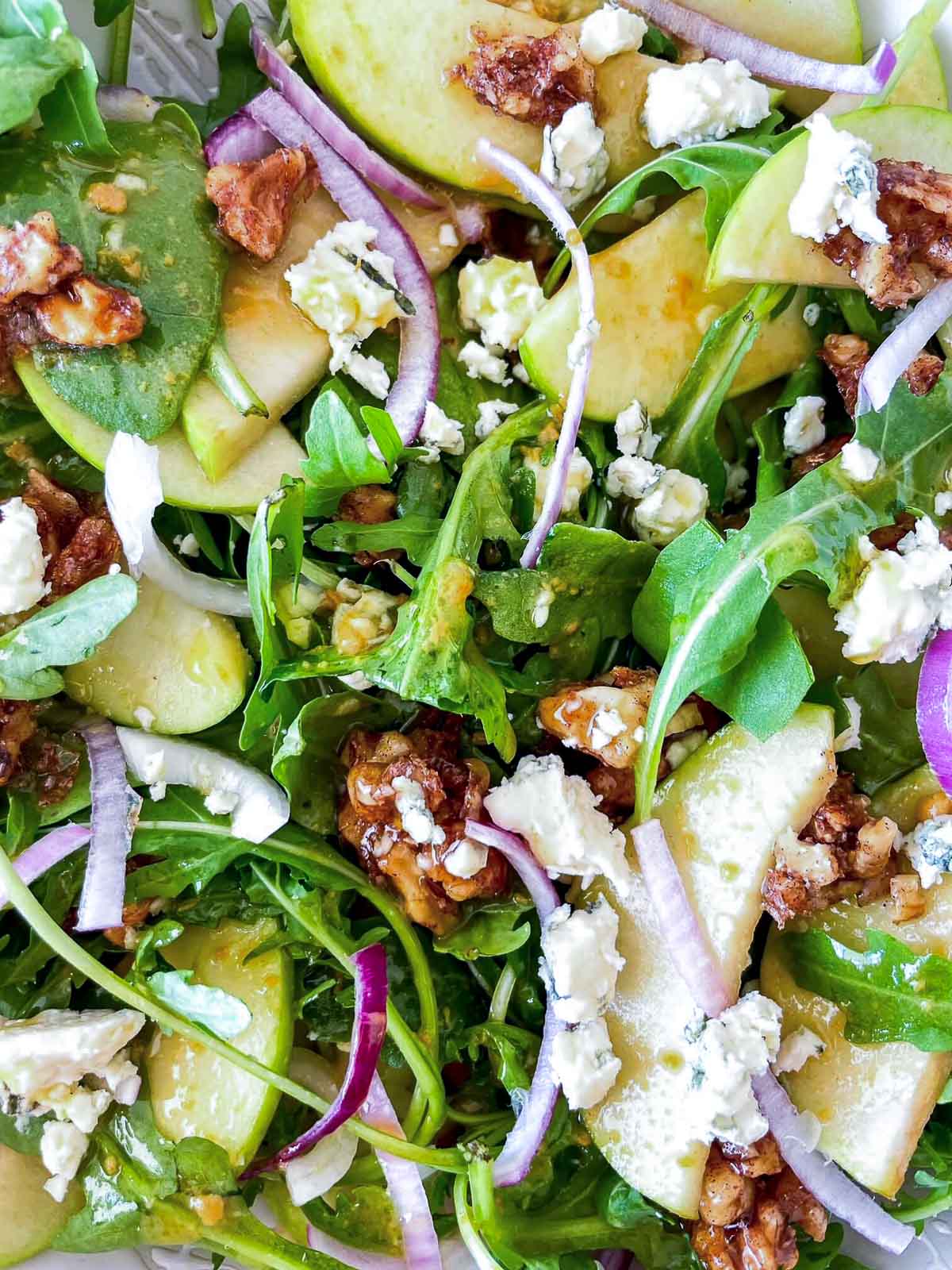 Close-up of arugula leaves, apple slices, blue cheese crumbles, and candied walnut pieces glistening with vinaigrette.