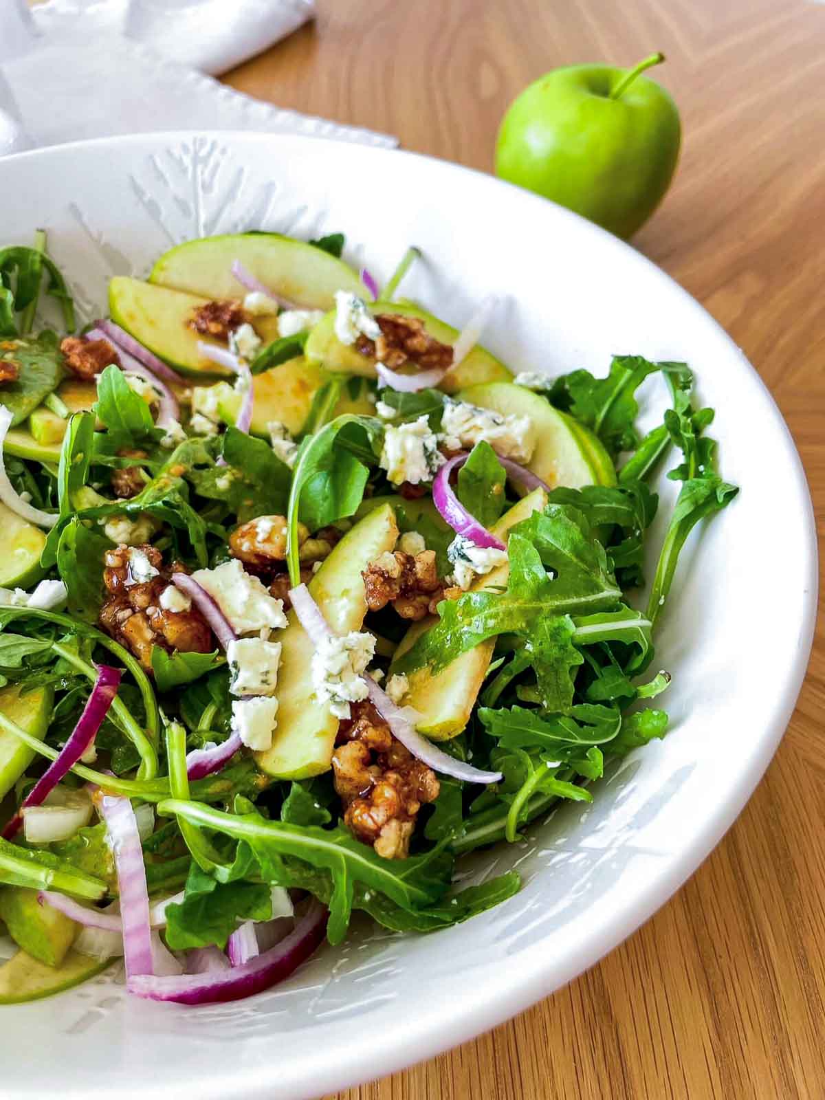 Side angle of apple walnut salad with white balsamic vinaigrette in a white bowl, with a green apple resting in the background.