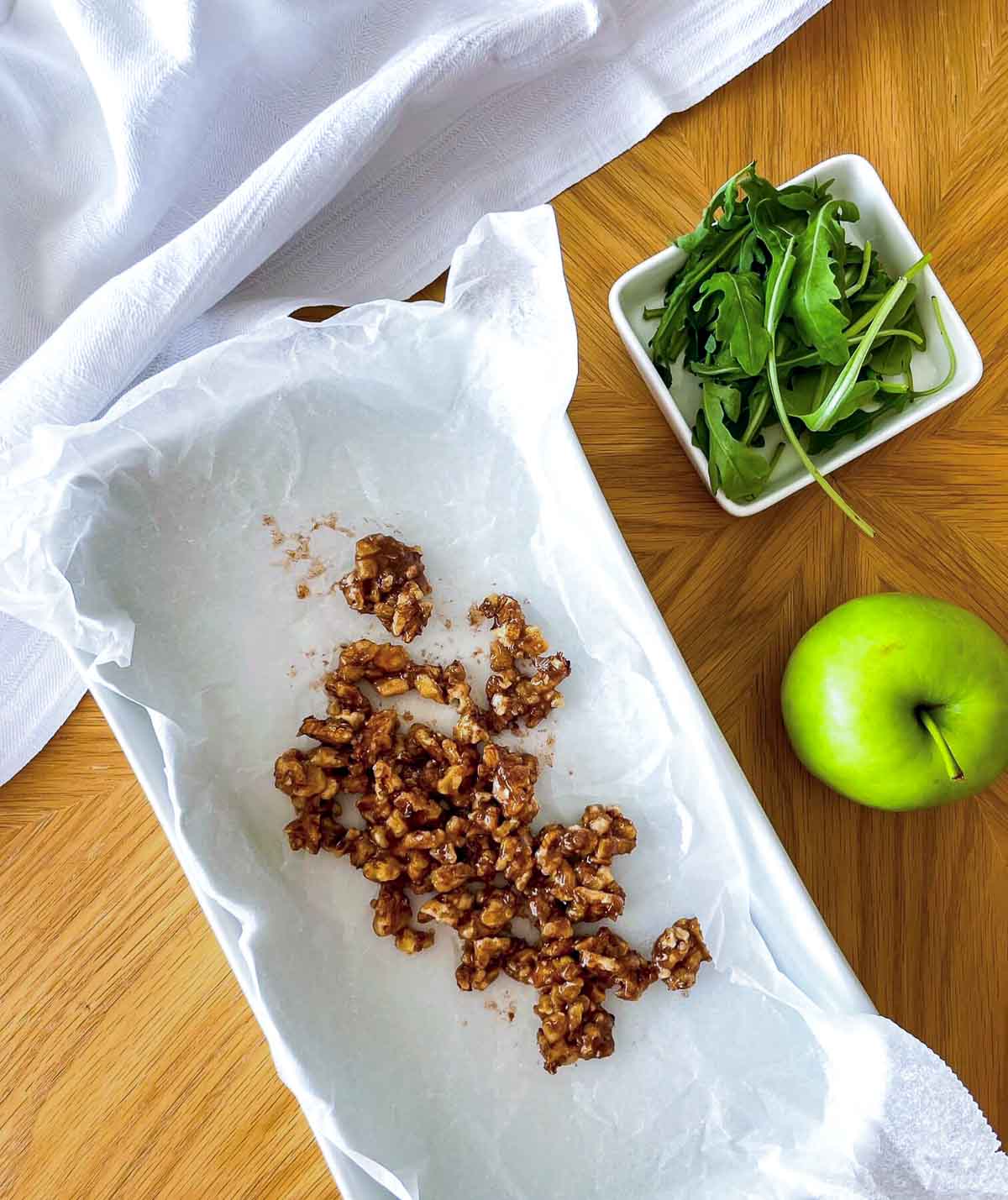 Candied walnuts cooling on parchment paper next to a bowl of arugula and a whole green apple.