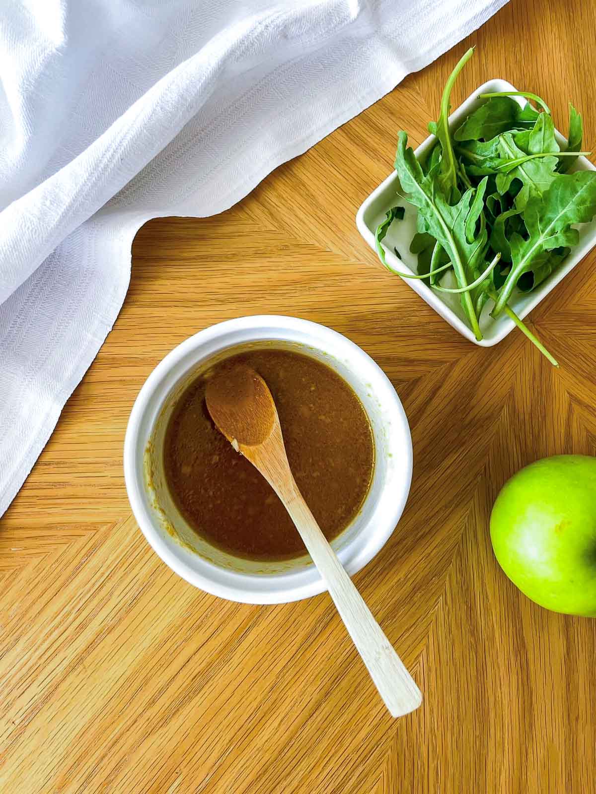 White balsamic vinaigrette in a small bowl with a wooden spoon, next to a bowl of arugula and a green apple.