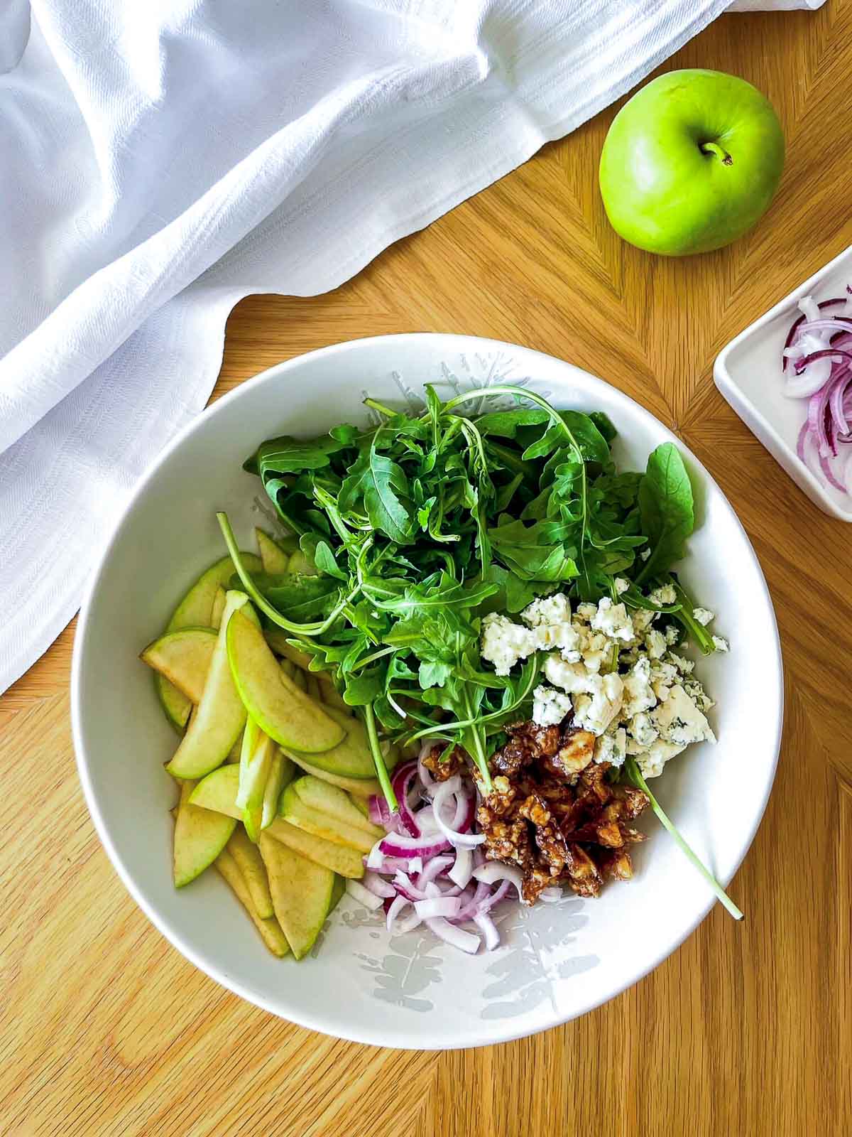 Salad bowl with arugula, sliced apples, red onion, blue cheese, and candied walnuts arranged in sections before tossing.