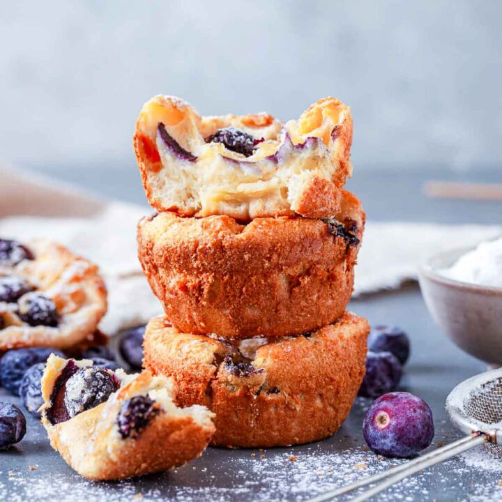 Two golden blueberry popovers stacked on top of each other, the top one torn open to show the light, airy inside studded with blueberries.