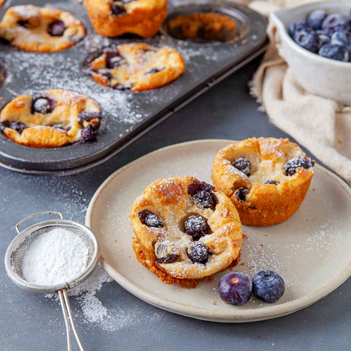 A muffin tin of baked blueberry popovers next to a plate with two popovers and a sifter of powdered sugar nearby.