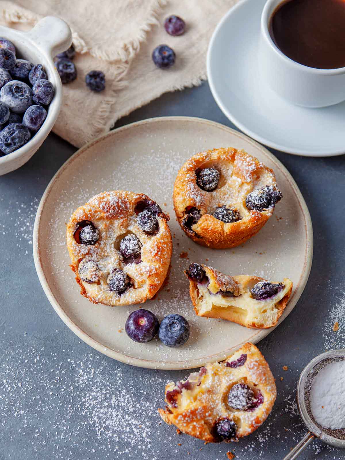 Two blueberry popovers on a ceramic plate dusted with powdered sugar, served with fresh blueberries and a cup of coffee.