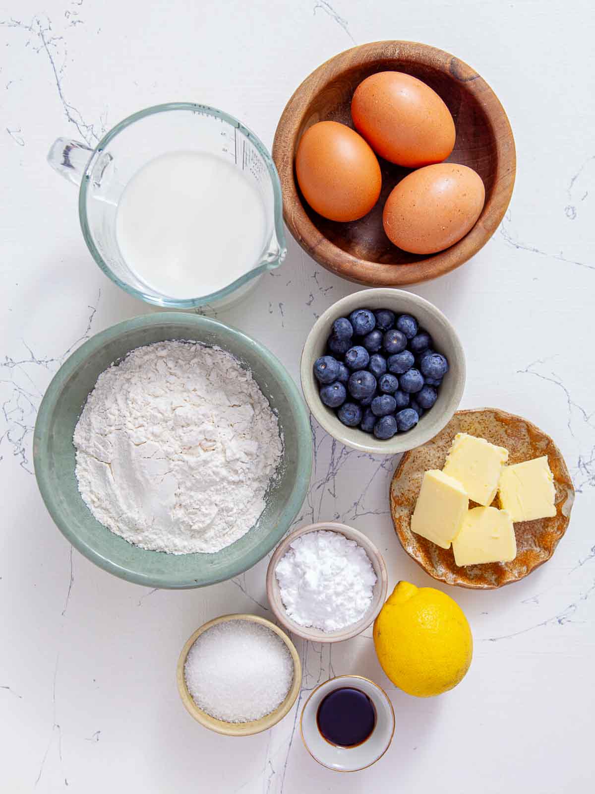 Ingredients are arranged on a marble counter, including a bowl of flour, three eggs, a pitcher of milk, fresh blueberries, cubed butter, a lemon, and small bowls of sugar and vanilla.