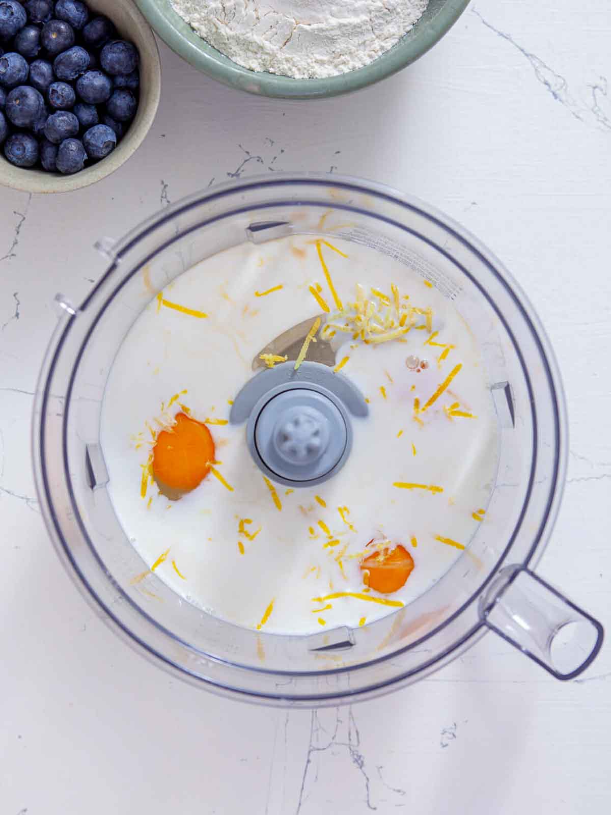 A food processor bowl holding milk, two cracked eggs, lemon zest, and vanilla extract, not yet blended.