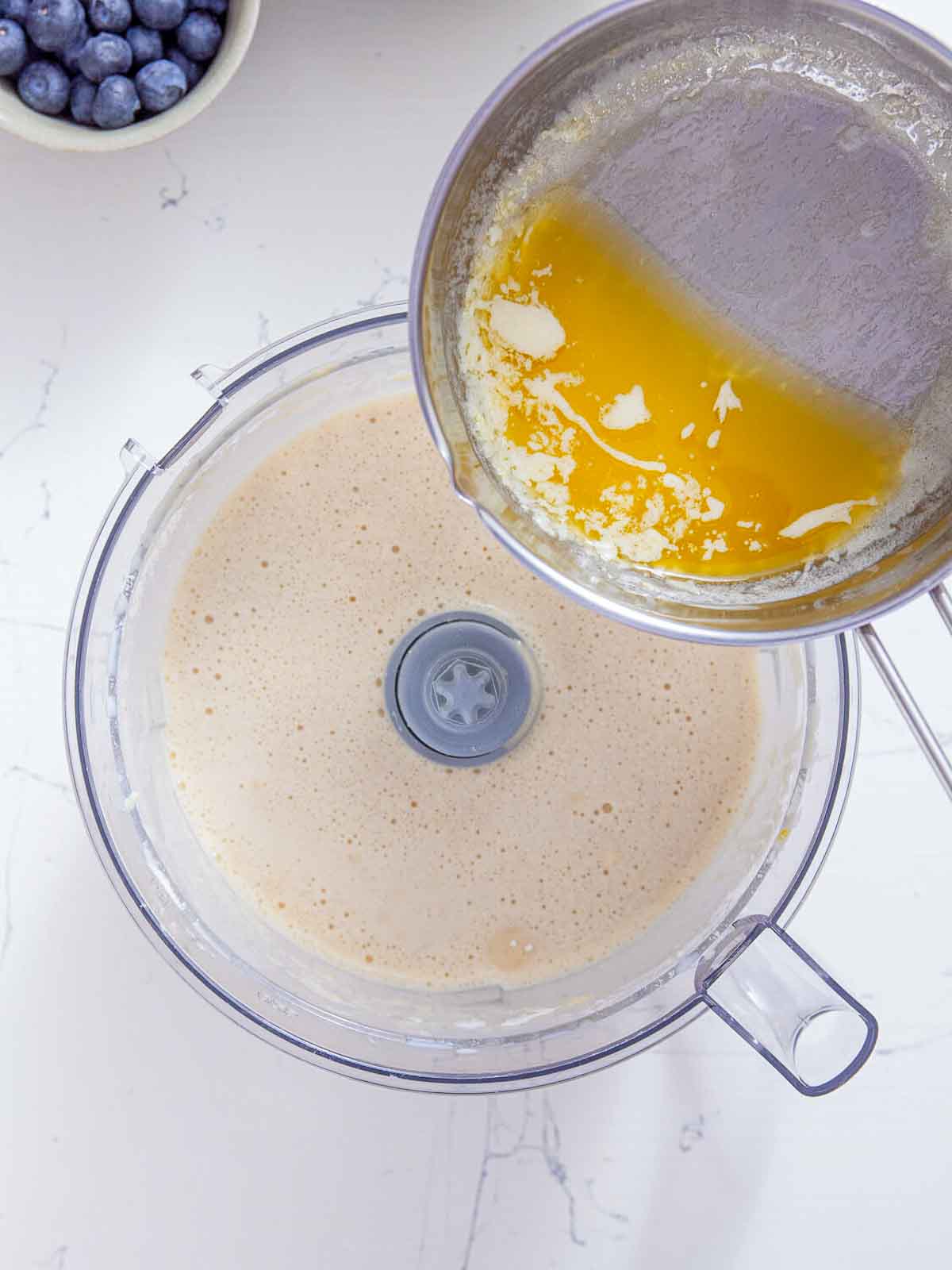 Melted butter being poured from a small saucepan into a food processor bowl of pale, smooth batter.