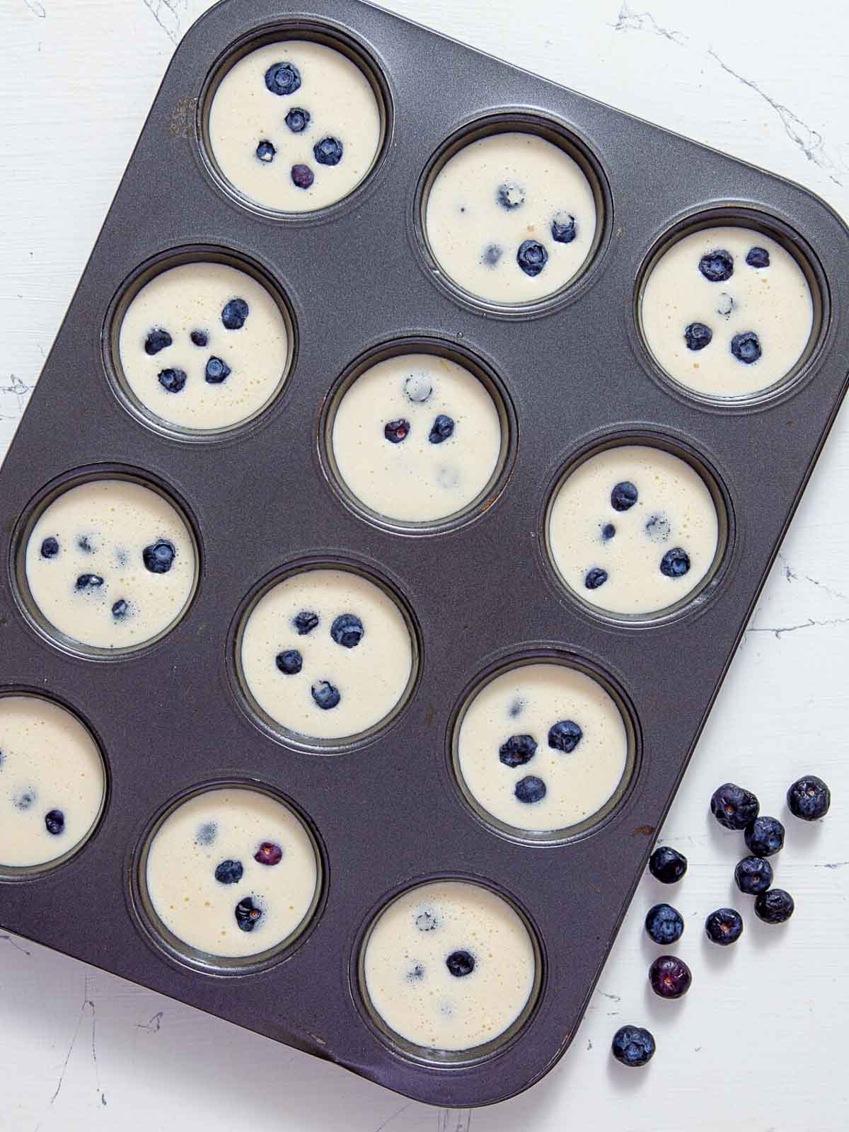 A muffin tin with each cup filled halfway with pale batter and topped with a few fresh blueberries, ready to go in the oven.