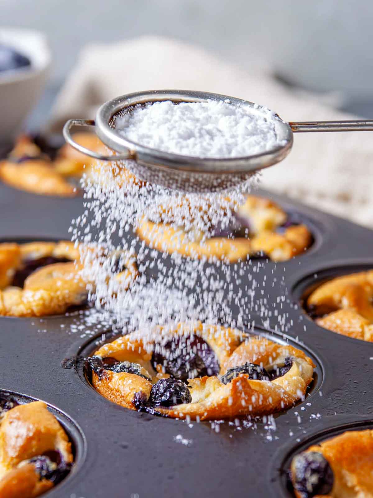 Powdered sugar being sifted through a small strainer over golden baked blueberry popovers still in the muffin tin.