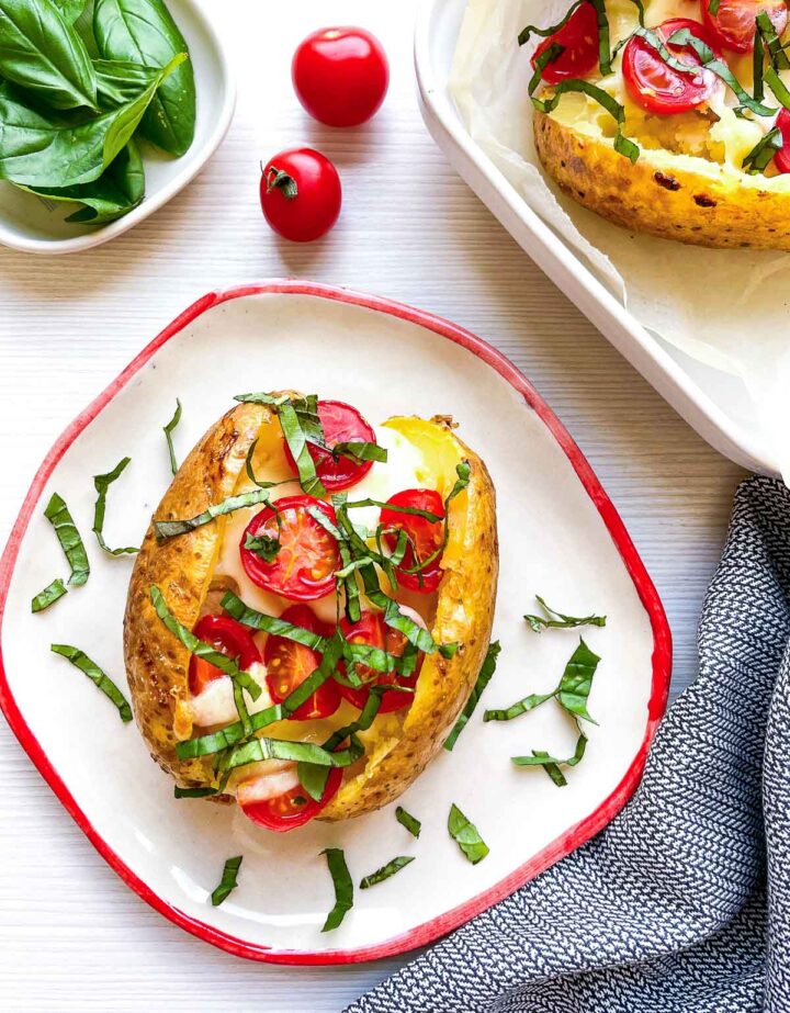 Overhead shot of a caprese baked potato on a red-rimmed plate, surrounded by fresh basil leaves, whole cherry tomatoes, and a striped napkin, with a second potato in a baking dish nearby.