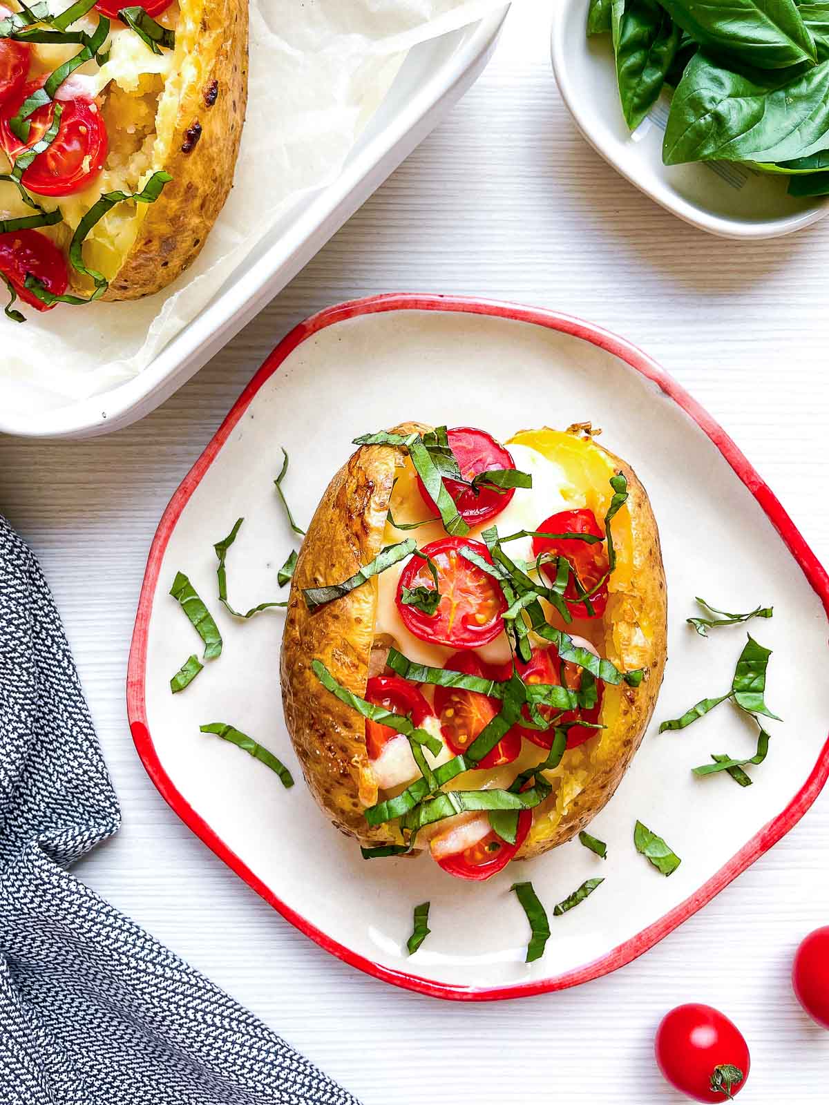 A finished caprese baked potato on a plate, with a second potato still in its parchment-lined baking dish in the background and fresh cherry tomatoes nearby.