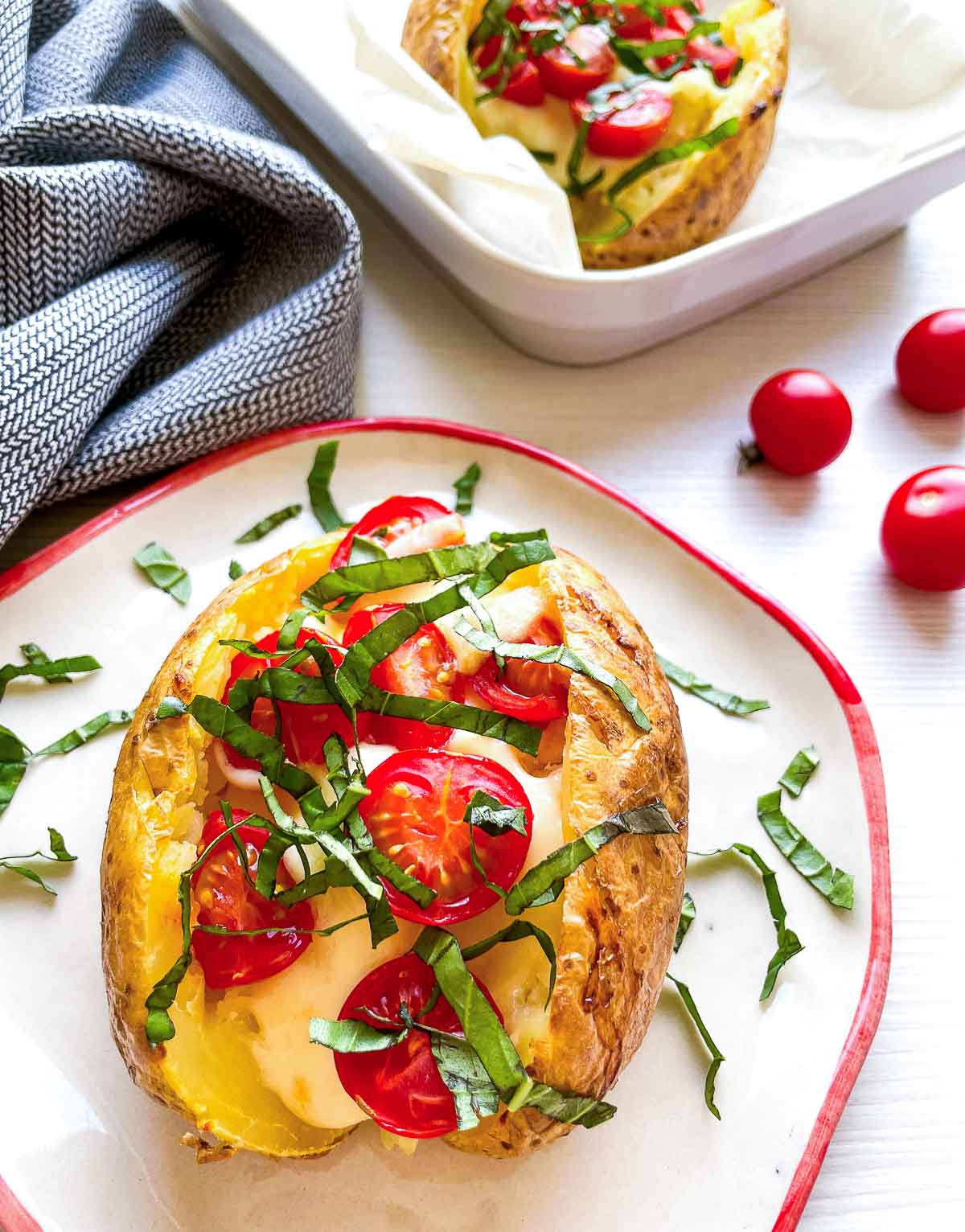 A caprese baked potato on a plate in the foreground, with a second potato in a parchment-lined baking dish and loose cherry tomatoes in the background.