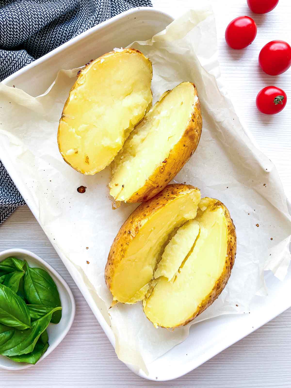 Two baked potatoes sliced in half on a parchment-lined tray, insides mashed with a fork, skins golden and unfilled.