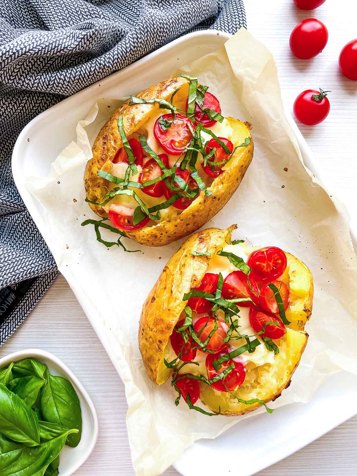 Two finished baked potatoes on a parchment-lined tray, topped with melted mozzarella, cherry tomatoes, and sliced fresh basil.