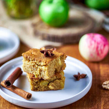 A stack of two cinnamon apple blondies on a white plate, garnished with star anise, next to a curled cinnamon stick and fresh green apples.