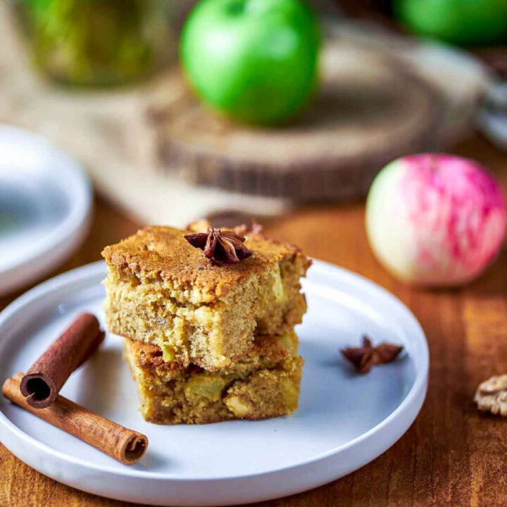 A stack of two cinnamon apple blondies on a white plate, garnished with star anise, next to a curled cinnamon stick and fresh green apples.