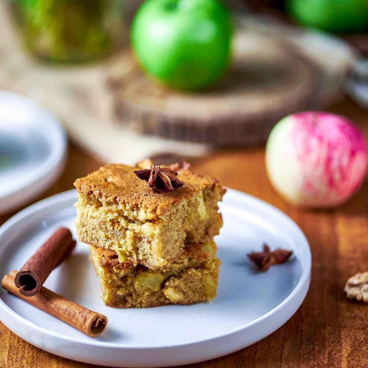 A stack of two cinnamon apple blondies on a white plate, garnished with star anise, next to a curled cinnamon stick and fresh green apples.
