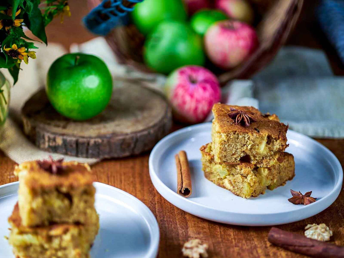 Two plates of stacked apple blondie squares on a wood table, with a basket of fresh apples and yellow flowers behind them