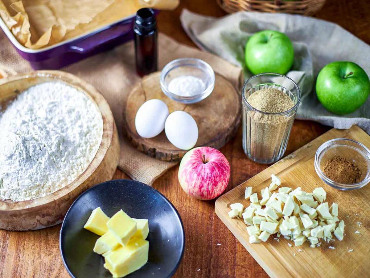 Overhead view of blondie ingredients including a bowl of flour, two eggs, cinnamon, baking powder, chopped white chocolate, cubed butter, and whole green and red apples.
