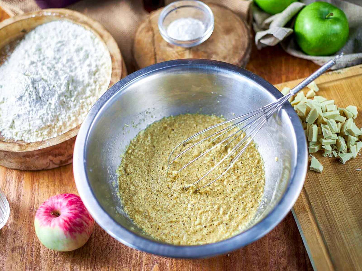 A whisk mixing a smooth, light brown batter in a metal bowl, with flour and an apple in the background.
